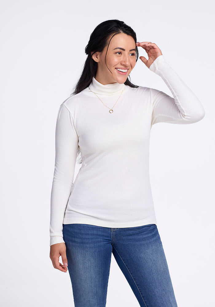 A smiling person with long dark hair leans to the side against a plain backdrop, one hand touching their head. They wear Woolx's Peyton Turtleneck in buttercream and blue jeans, complemented by a small necklace.