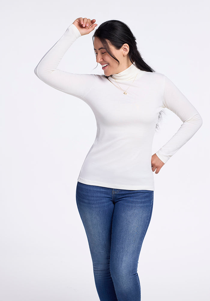 A person with long dark hair smiles, posing with one arm bent overhead. They're wearing Woolx's buttery Peyton Turtleneck and blue jeans against a light background.