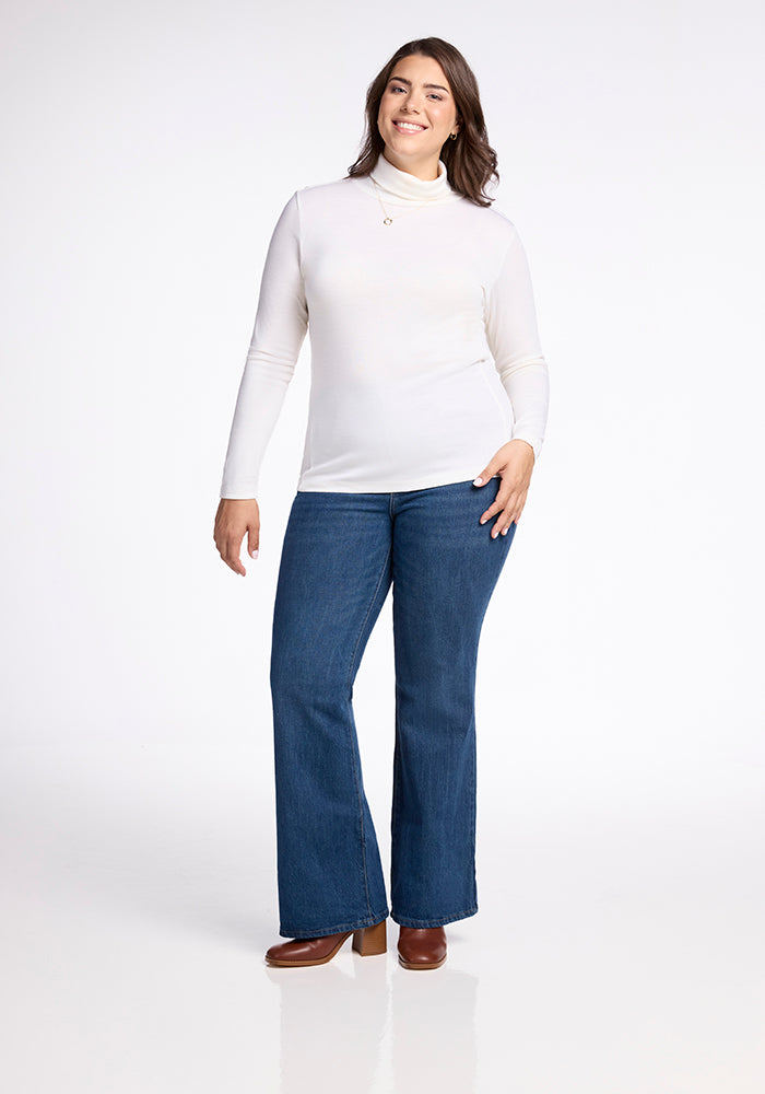 A person with long brown hair smiles in a Woolx Peyton Turtleneck in Buttercream and blue flared jeans, stylishly posing with one hand on their hip and wearing brown shoes against a plain white background.