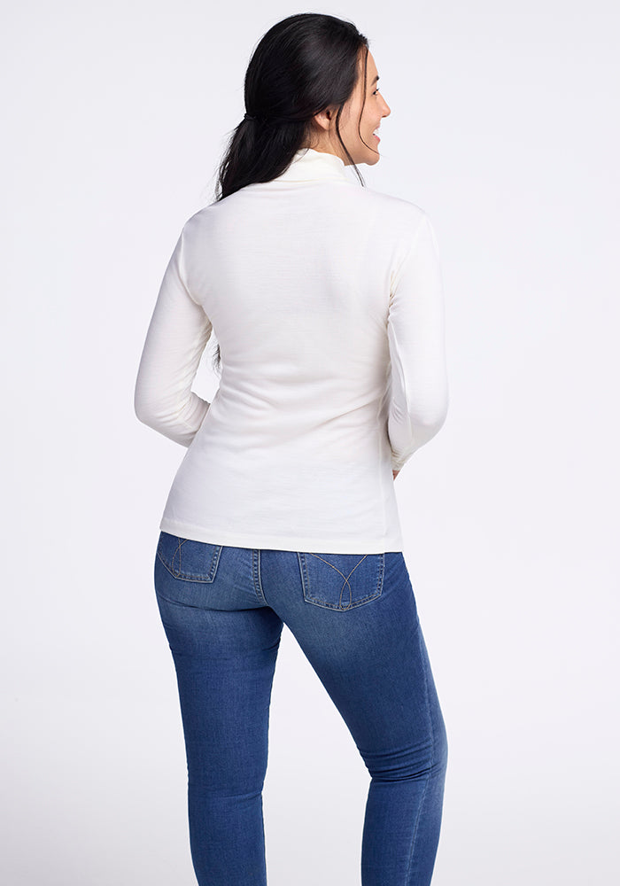 A person with long dark hair faces away, featuring the simplicity of the Woolx Peyton Turtleneck in Buttercream with blue jeans. The background is plain white, highlighting the breathable fabric's elegance.