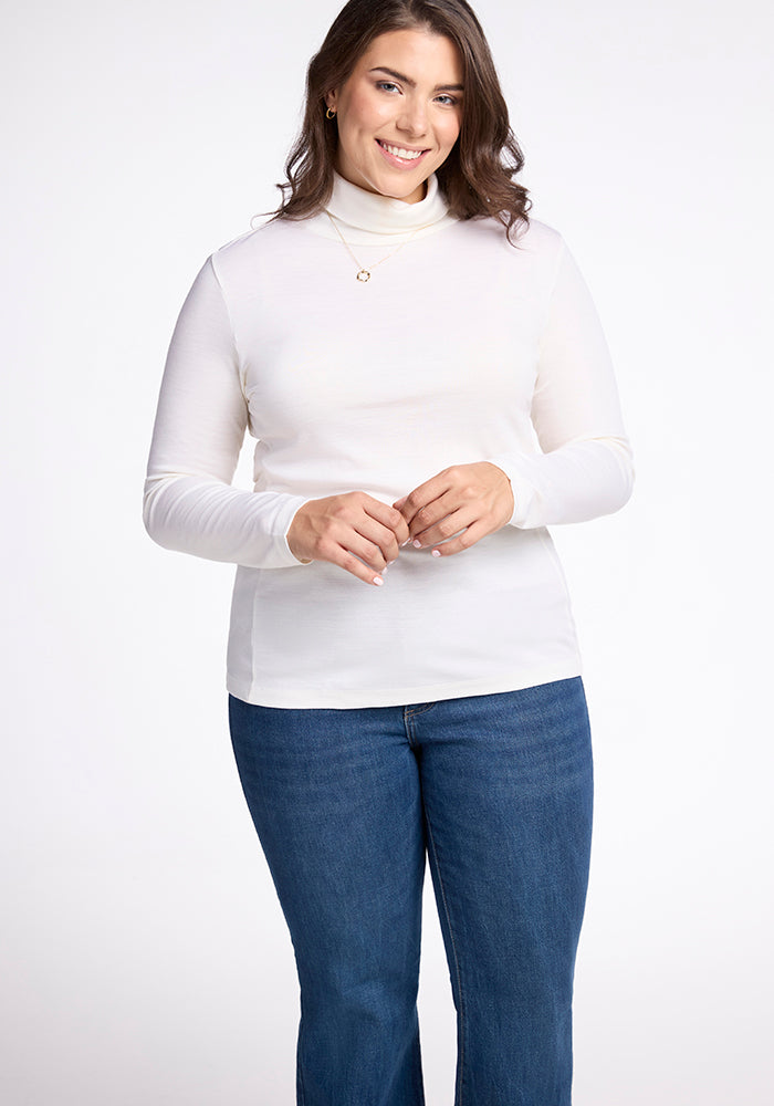 A smiling woman in a Woolx Peyton Turtleneck, buttercream shade, stands against a plain backdrop. She accessorizes her long brown hair with a small necklace.