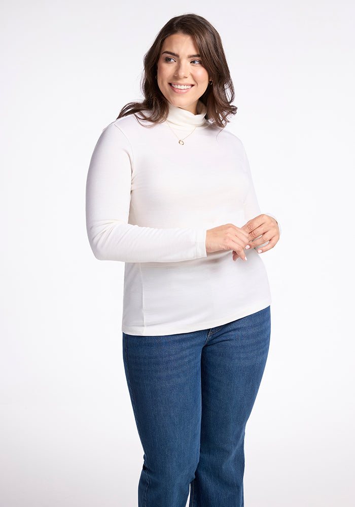 A person with long hair smiles against a plain background, wearing the Buttercream Peyton Turtleneck from Woolx made with breathable Merino wool, along with blue jeans. Their hands are gently clasped in front of them. 