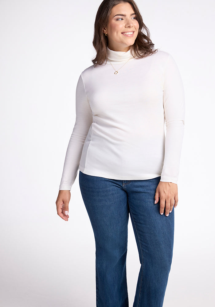 A woman wearing the Woolx Peyton Turtleneck in Buttercream and blue jeans stands against a plain white background, smiling and gazing slightly to the side.