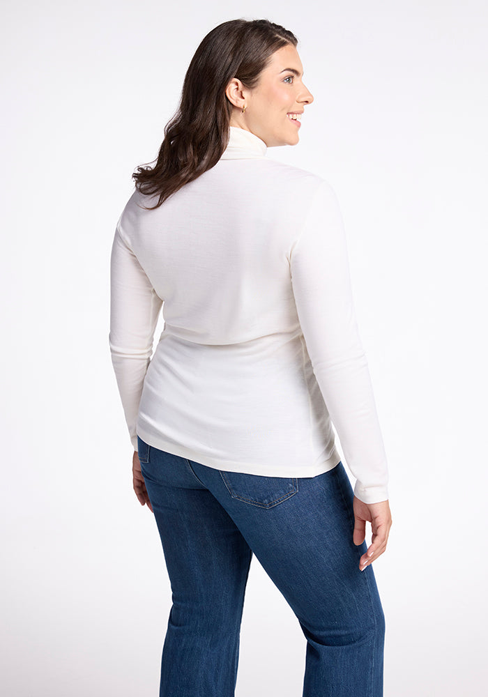 A person with long brown hair smiles while standing sideways, slightly facing left. They're wearing a Buttercream Peyton Turtleneck by Woolx and blue jeans against a plain white background.