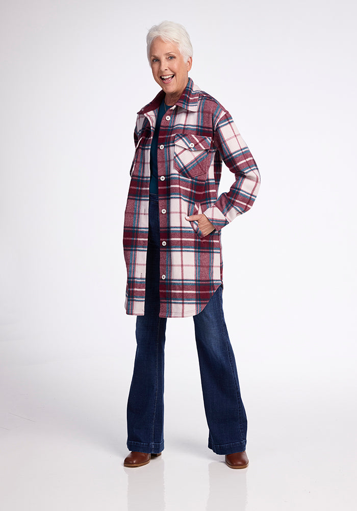 An older woman with short white hair smiles while posing in the Woolx Sutton Shacket - Frozen Cranberry Plaid over a dark shirt, blue jeans, and brown shoes. She stands against a white background with one hand in her shacket pocket.