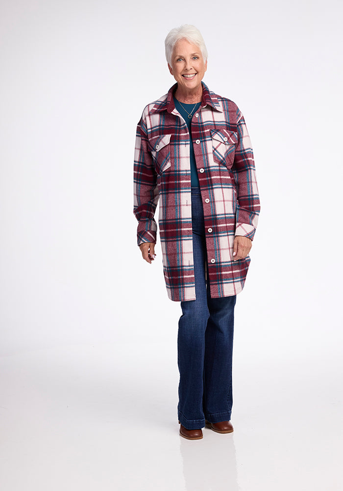 A smiling older woman with short gray hair models the Woolx Sutton Shacket in Frozen Cranberry Plaid, paired with blue jeans and brown shoes, standing against a plain white background.
