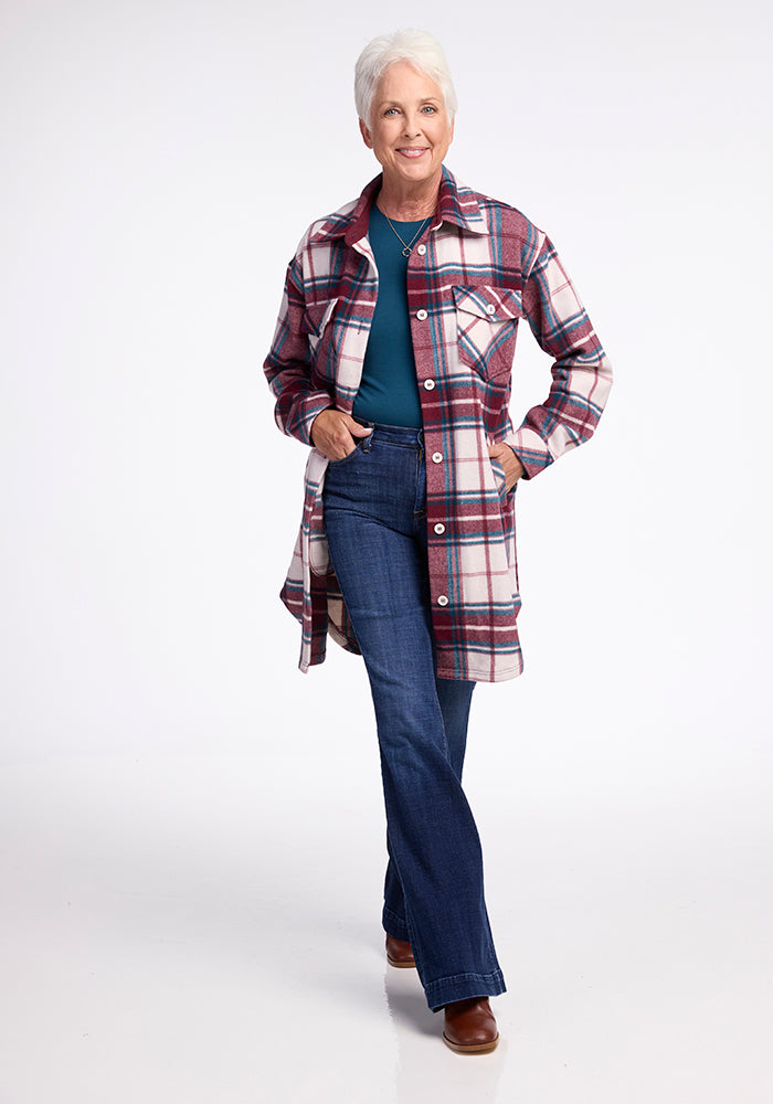 A smiling older woman with short white hair wears the Woolx Sutton Shacket in Frozen Cranberry Plaid, paired with a teal shirt, blue jeans, and brown shoes. She stands with one hand in her pocket against a white background.
