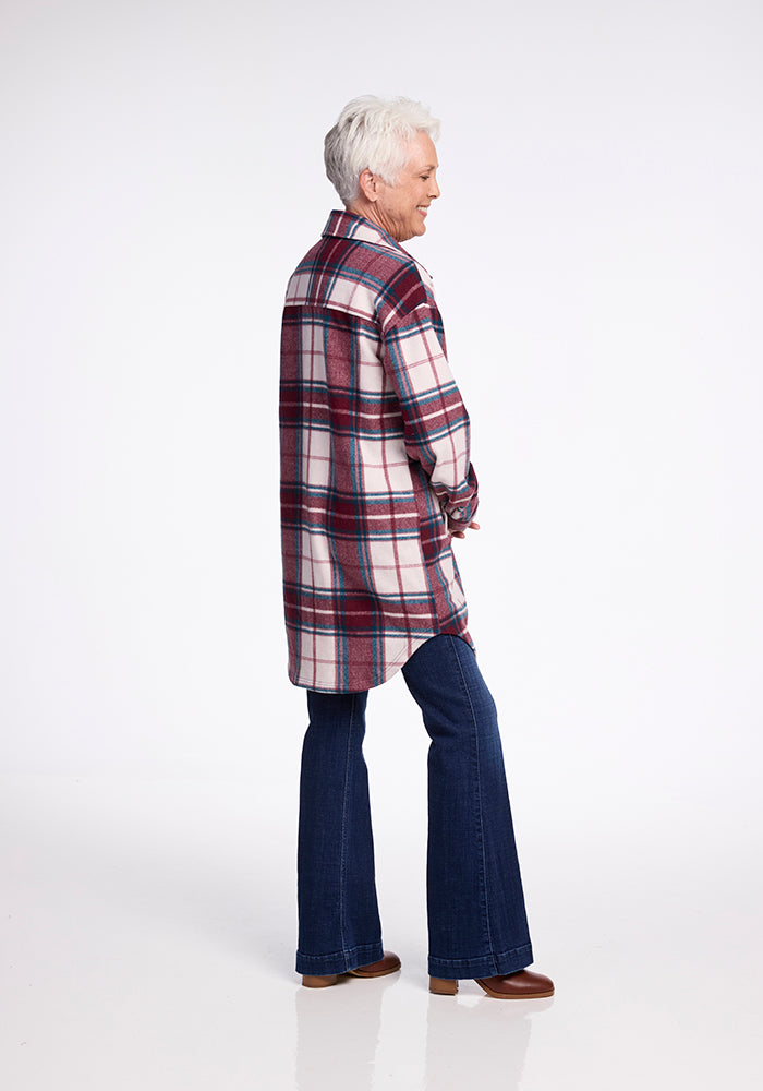 An older woman with short white hair stands sideways, smiling. She wears the Woolx Sutton Shacket in Frozen Cranberry Plaid, flared blue jeans, and brown shoes against a plain white background.