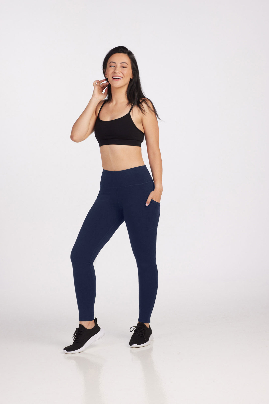A woman stands smiling against a white background, wearing a black sports bra, Woolx Piper Pocket Leggings in Deep Navy, and black athletic shoes.