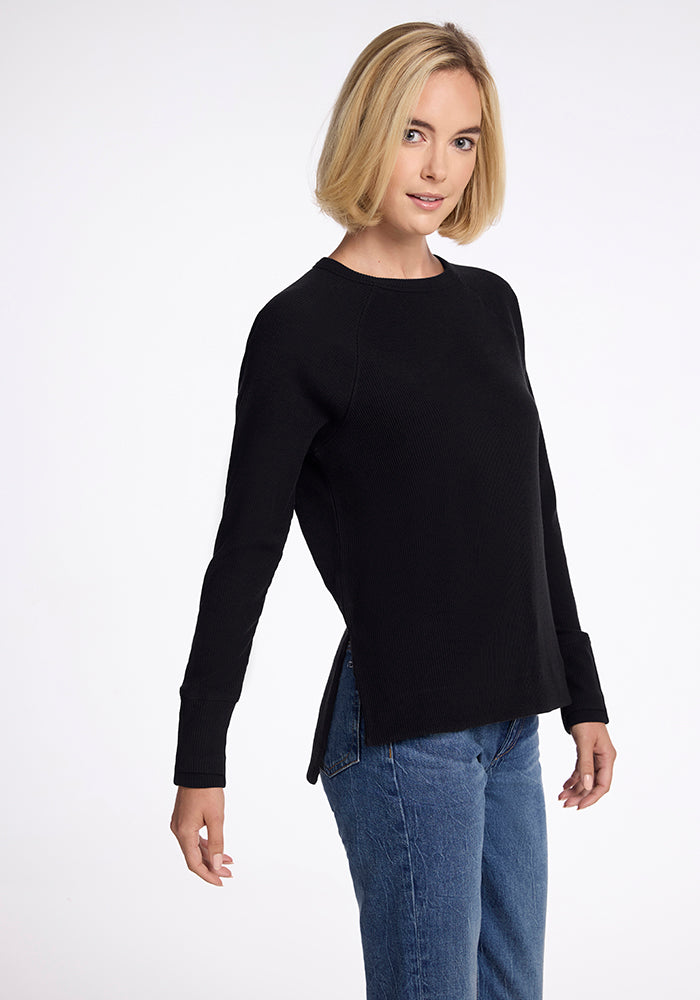Blonde woman wears Woolx Ainsley Petite Sweater in black and blue jeans, standing against a white background.