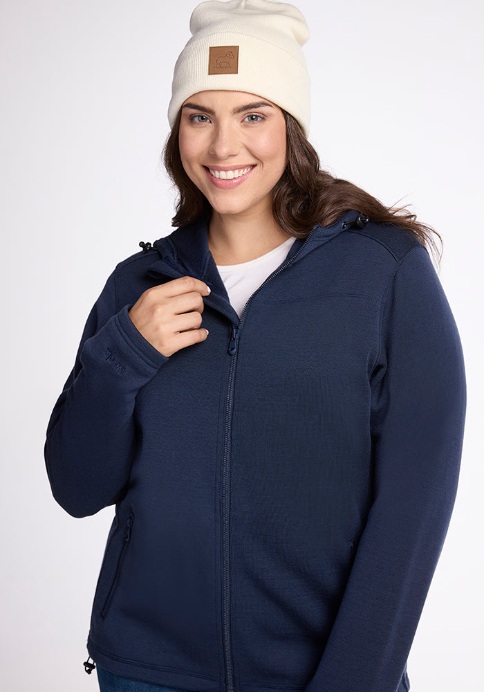 A person in a Woolx Cubby Hooded Sweatshirt in Deep Navy and a white beanie smiles while posing against a neutral background, gently adjusting the sweatshirt's zipper.