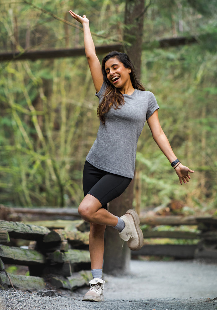 A joyful woman stands on a forest trail, playfully raising one arm and leg. She wears the Woolx Addie Crew Neck in Graphite Heather, black shorts, and hiking boots, surrounded by trees and wooden fences.
