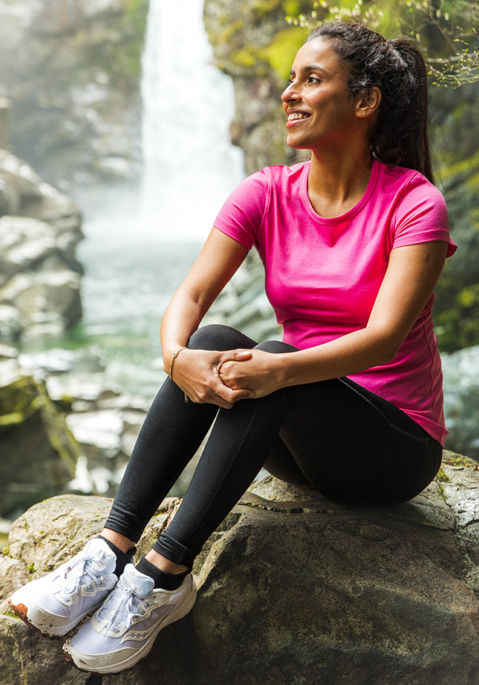 A woman in the Woolx Addie Crew Neck - Raspberry Fizz and black leggings sits on a rock, smiling amid a waterfall and lush greenery—enjoying an odor-free adventure.
