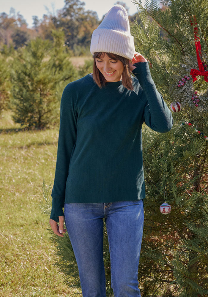 A woman wearing a white knit beanie, Woolx Ainsley Sweater in Deep Timber, and blue jeans stands outdoors by a decorated Christmas tree, smiling while looking down and touching her hat.
