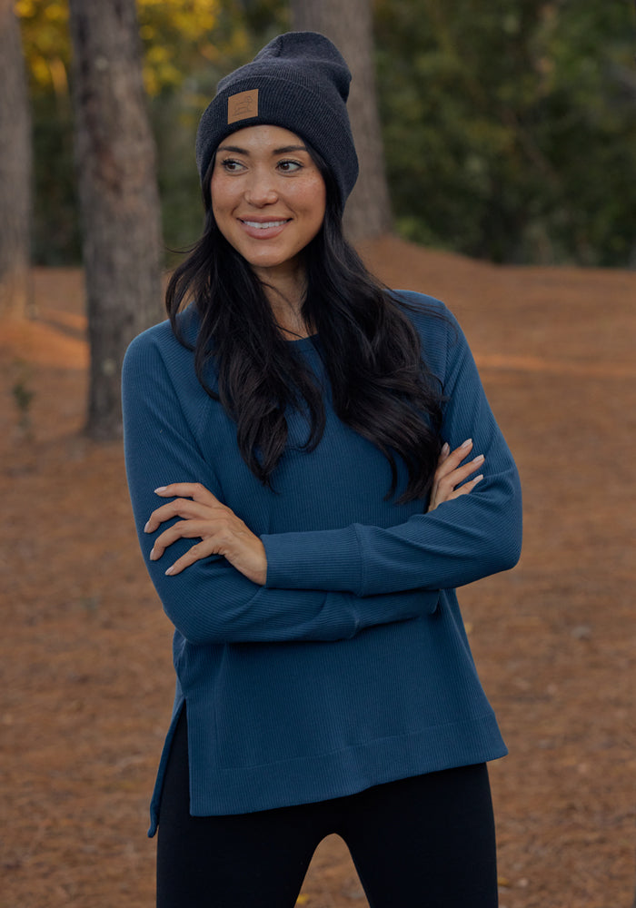 A woman with long dark hair, wearing a dark beanie, black pants, and a blue Woolx Ainsley Sweater in Deep Timber stands outdoors smiling with arms crossed amid tall trees and brown pine needles on the ground.