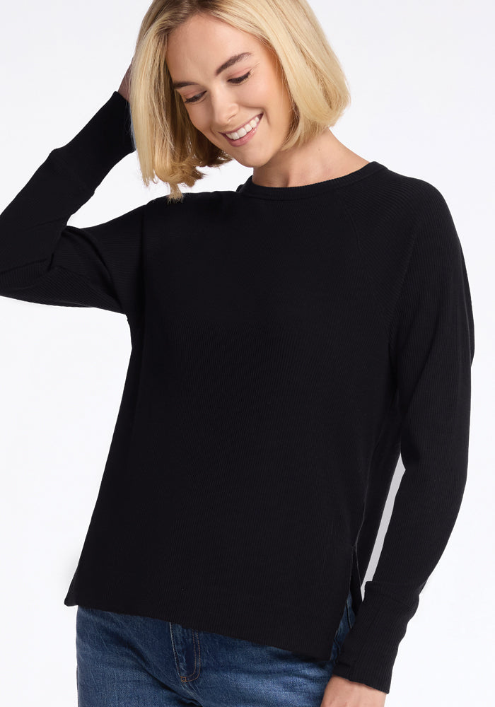 Smiling woman with short blonde hair wears the Woolx Ainsley Petite Sweater in black and blue jeans, looking down.