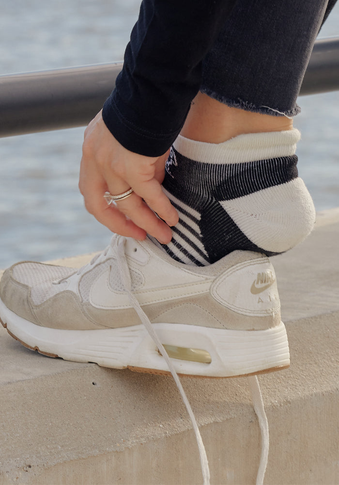 A person wearing white Nike sneakers with untied laces adjusts their Woolx Womens Amelia No Show Socks - Tuxedo Stripe while sitting on a concrete surface near water.