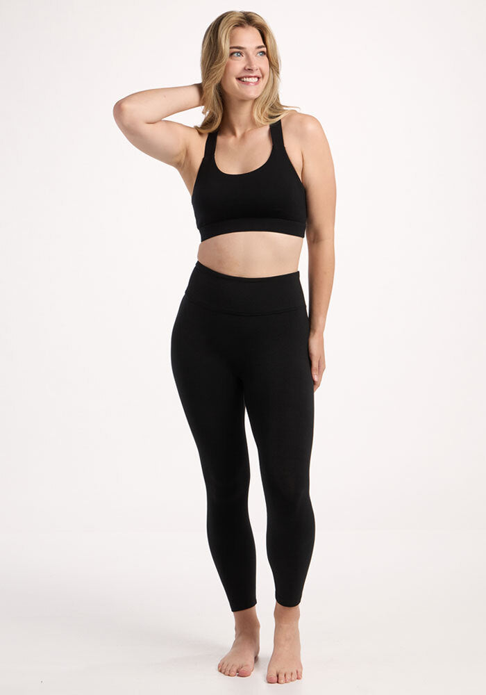 A woman in Woolx Angela Leggings - Black and a sports bra stands barefoot, smiling, with one hand in her hair.