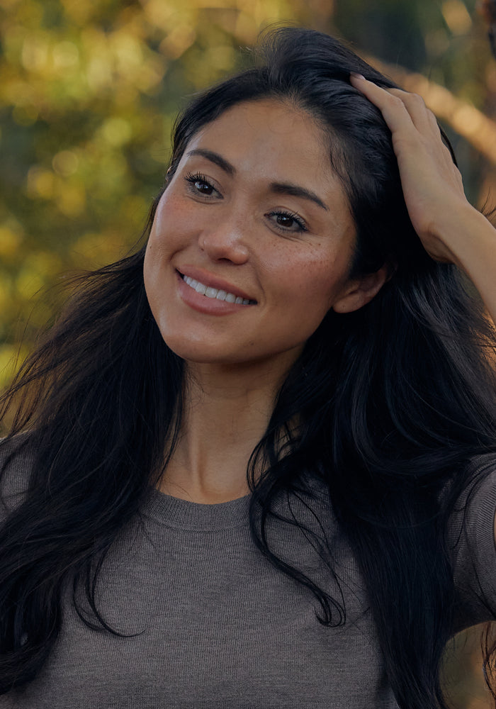 A woman with long dark hair smiles gently, touching her hair as she stands outdoors in a softly blurred green and yellow setting, wearing the Woolx Anna Crew Sweater in Simply Taupe.