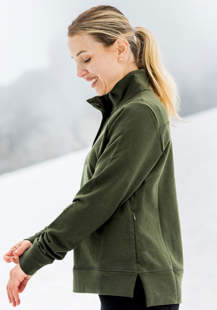 Woman in Woolx Annie 3 Button Pullover - Duffel Bag smiles outdoors on a snowy day, looking down at her sleeve.
