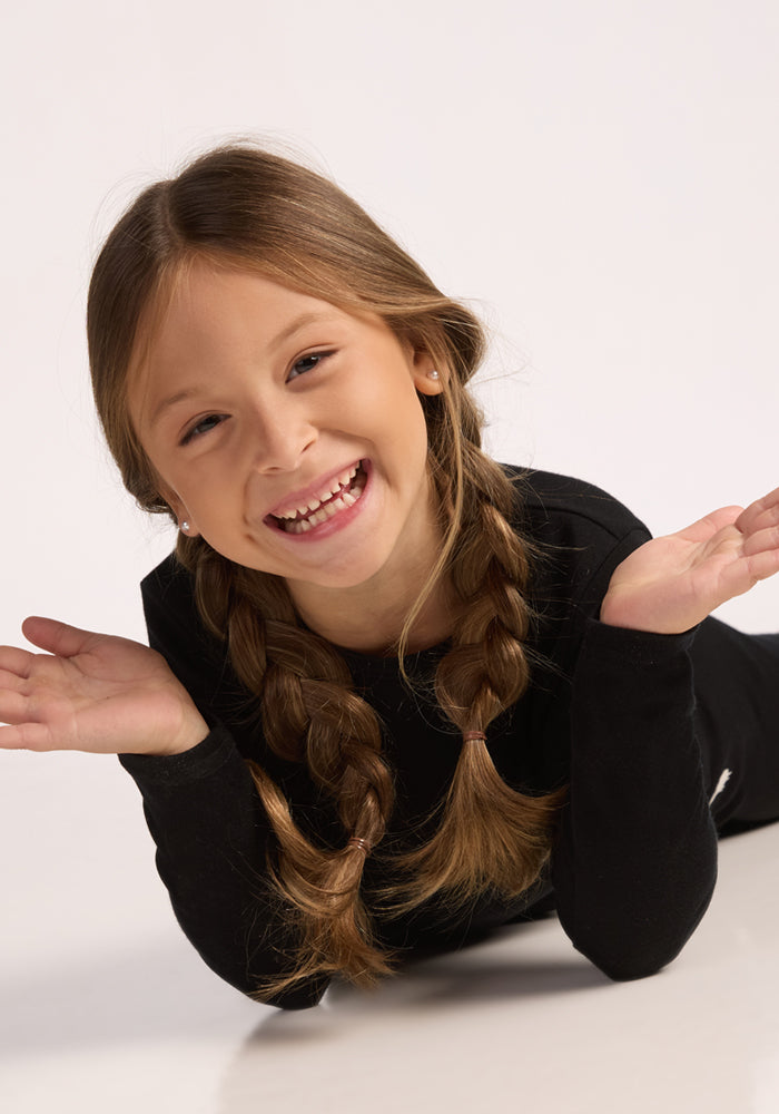 A smiling young girl with long brown hair in two braids lies on her stomach, playfully raising her hands. She wears the Woolx Kids Explorer Base Layer Top in black against a plain, light background.