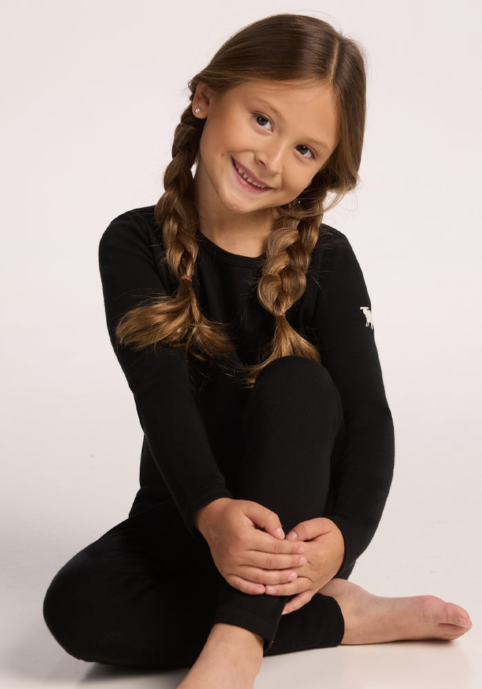 A young girl with long brown braids sits barefoot on the floor, smiling at the camera while wearing the Woolx Kids Explorer Base Layer Top in black against a plain light background.