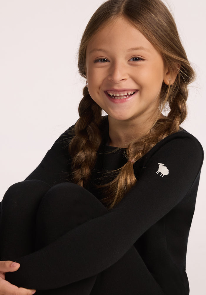 A smiling young girl with long brown hair in braids wears the Woolx Kids Explorer Base Layer Top in black, featuring a small white sheep logo on the shoulder, as she sits with her knees up against a plain light background.