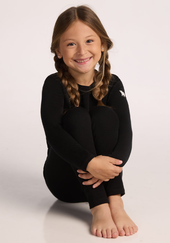 A young girl with long brown hair in braids sits barefoot, hugging her knees and smiling, wearing the Woolx Kids Explorer Base Layer Top in black with a small white animal logo on the shoulder.