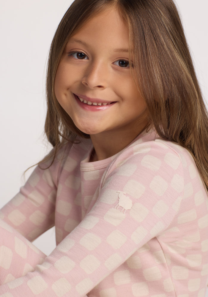 A young girl with long brown hair smiles at the camera, wearing the Woolx Kids Explorer Base Layer Top in Hopscotch Pink—a light pink, moisture-wicking, long-sleeve Australian Merino wool shirt featuring a white checkered pattern and a small animal logo on the sleeve.