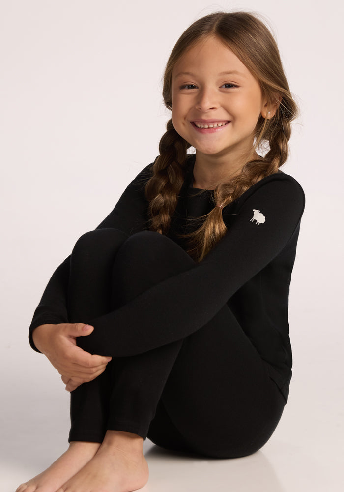A smiling young girl with long brown braided hair sits barefoot, wearing Woolx Kids Explorer Base Layer Bottoms in black—warm, cozy merino wool perfect for chilly days.