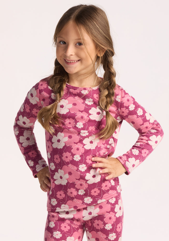 A young girl with long brown braided hair stands smiling with hands on hips, wearing the Woolx Kids Explorer Base Layer Top in Rosie Posie—a pink Australian Merino wool top with a white and magenta floral pattern. The background is plain and light-colored.
