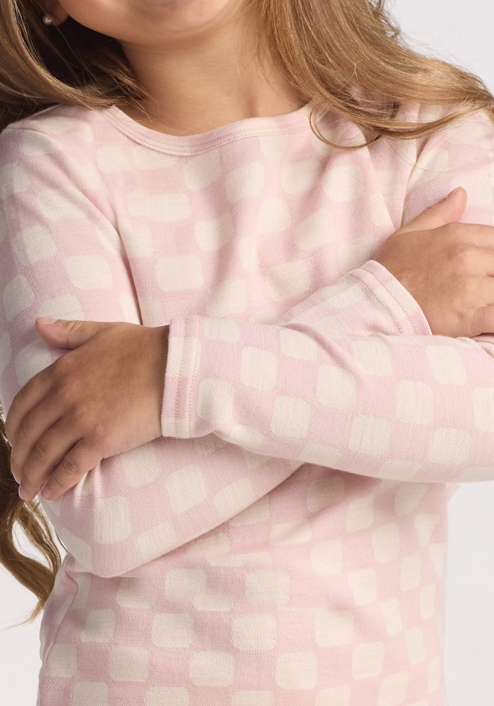 A child with long light brown hair stands with arms crossed, wearing a pink and white checkered shirt over the Woolx Kids Explorer Base Layer Top in Hopscotch Pink, showcasing relaxed, moisture-wicking comfort.