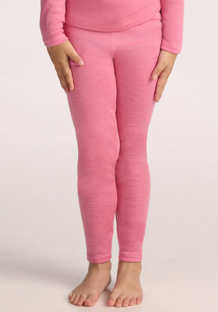 A child stands barefoot on a light surface, wearing a bright pink long-sleeve top and Woolx Kids Explorer Base Layer Bottoms in Pinky Promise. Only the body from shoulders to feet is visible, with relaxed arms and one hand gently touching the thigh. 