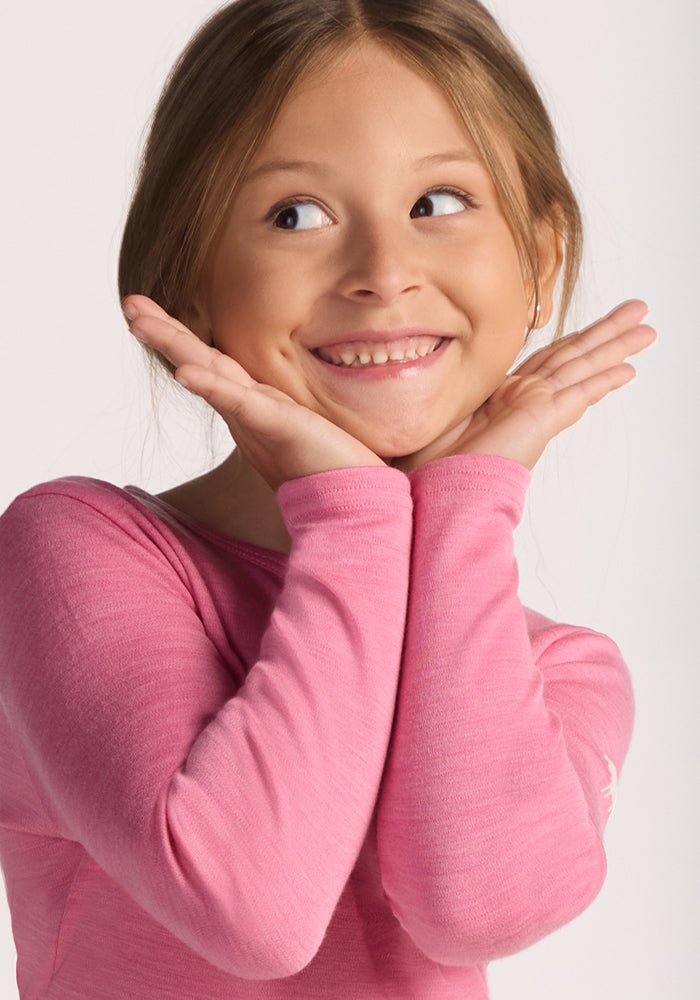 A smiling young girl with light brown hair looks to the side, resting her face in her hands. She wears the Woolx Kids Explorer Base Layer Top in Pinky Promise, appearing happy and playful against a plain background.