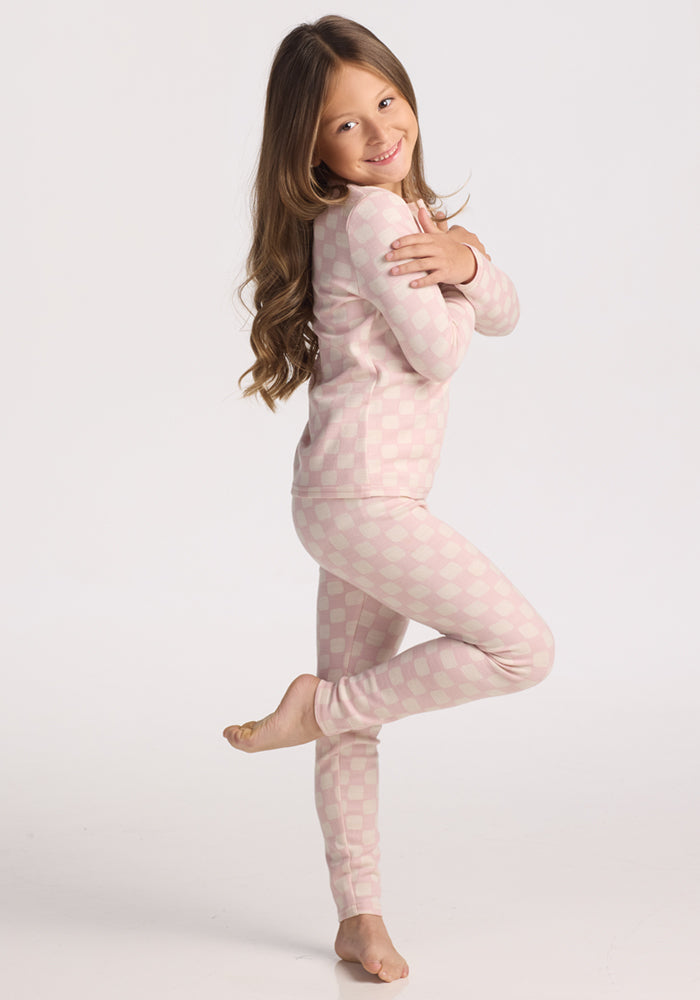 A young girl with long brown hair stands on one foot, smiling and hugging herself. She is wearing Woolx Kids Explorer Base Layer Bottoms in Hopscotch Pink against a plain white background.