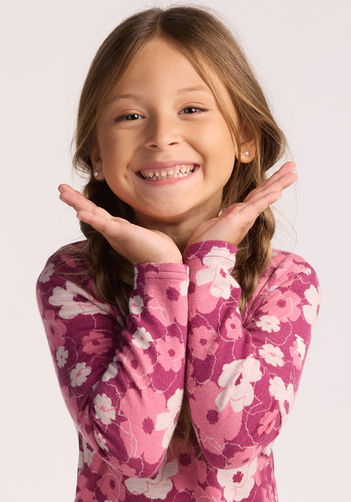 A young girl with long brown hair in braids smiles brightly, wearing the Woolx Kids Explorer Base Layer Top - Rosie Posie. This warm, soft Australian Merino wool shirt features pink and purple florals against a light background.