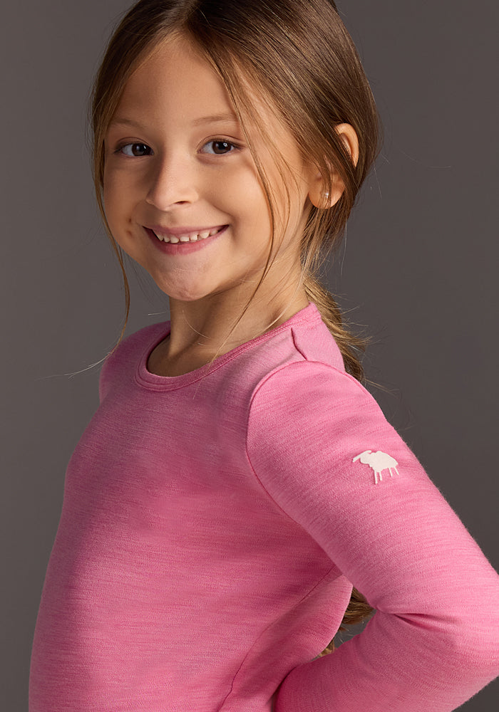A smiling young girl with long brown hair in a ponytail wears the Woolx Kids Explorer Base Layer Top - Pinky Promise, featuring a small white sheep logo on the sleeve, standing against a plain gray background.