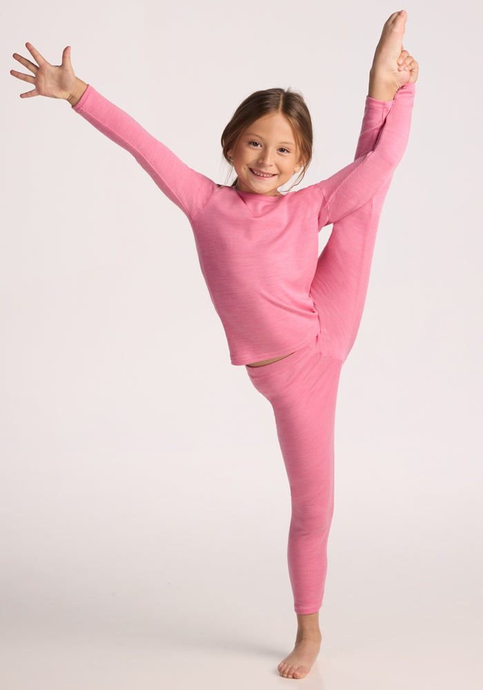 A young girl in Woolx Kids Explorer Base Layer Bottoms - Pinky Promise performs a standing split, smiling confidently as she shows off her stylish and warm merino leggings against a simple, light background.