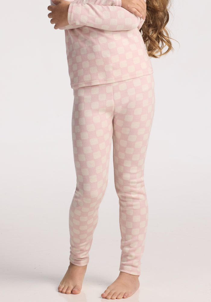 A child stands barefoot with arms crossed, facing slightly to the side against a white background, wearing Woolx Kids Explorer Base Layer Bottoms in Hopscotch Pink. The child’s face is not visible.