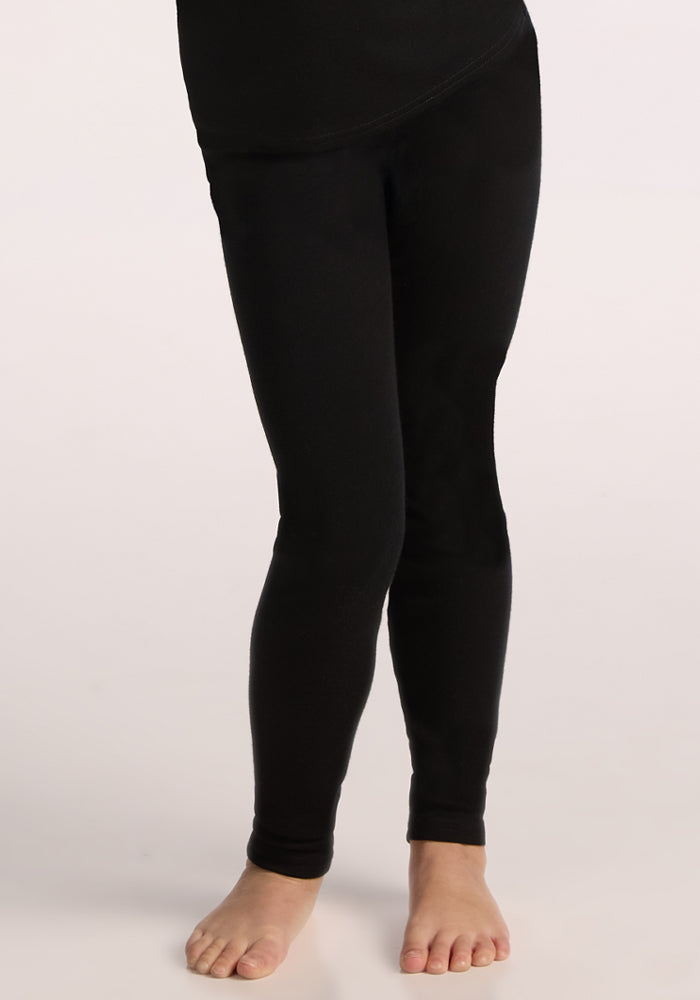 A child wears Woolx Kids Explorer Base Layer Bottoms in black, standing barefoot on a light surface, with only the lower body visible from the waist down.