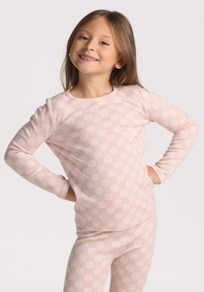A young child with long brown hair smiles with hands on hips, wearing the Woolx Kids Explorer Base Layer Top in Hopscotch Pink, standing against a plain white background. 
