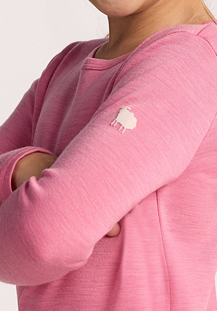 A girl in a Woolx Kids Explorer Base Layer Top - Pinky Promise stands with arms crossed. A small white sheep icon decorates the left sleeve, highlighting the soft Australian Merino wool fabric.