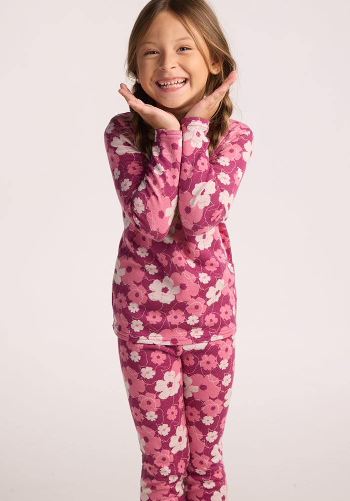 A young girl smiles cheerfully, hands framing her face, wearing Woolx Kids Explorer Base Layer Bottoms - Rosie Posie with matching pink and purple floral pajamas, standing against a plain light background.