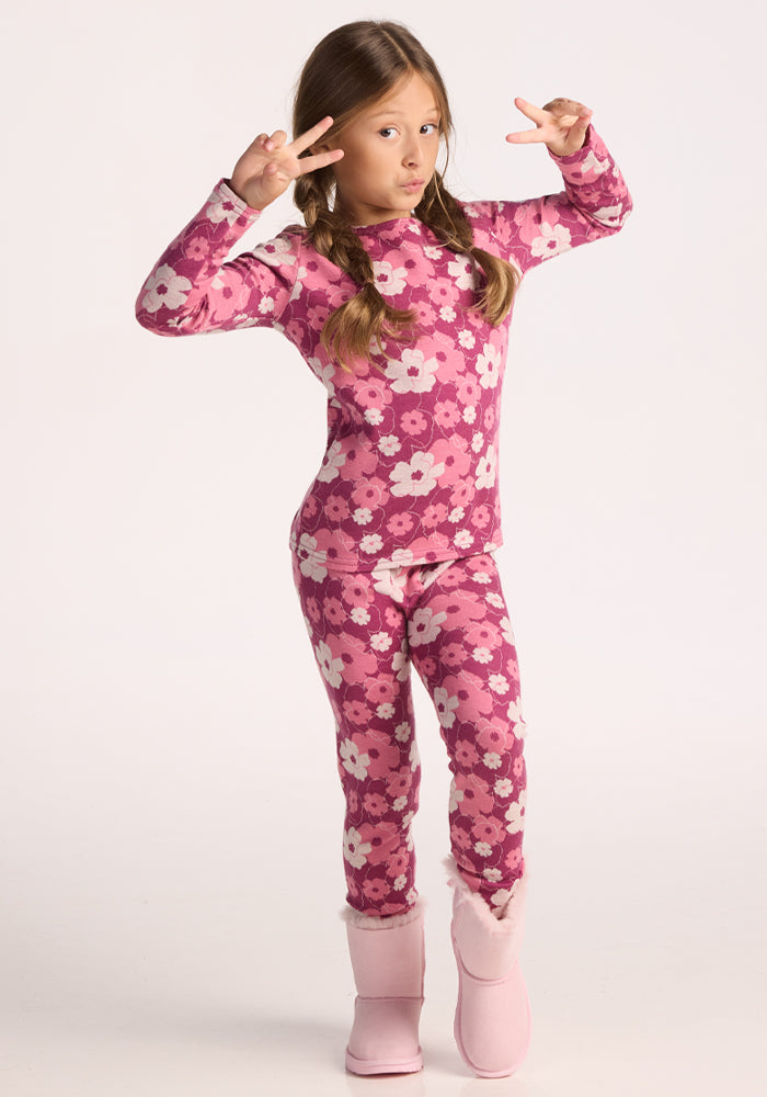 A young girl with long brown hair in pigtails makes peace signs, wearing Woolx's Kids Explorer Base Layer Top - Rosie Posie under a pink floral pajama set and pink fuzzy boots, standing against a light background.