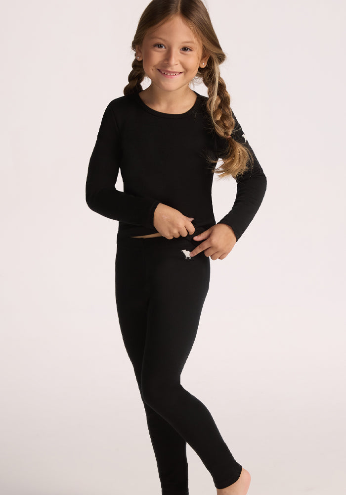 A young child with long braided hair, wearing a black long-sleeve top and Woolx Kids Explorer Base Layer Bottoms in black, smiles and lifts the top to reveal the waistband, standing barefoot against a plain light background.