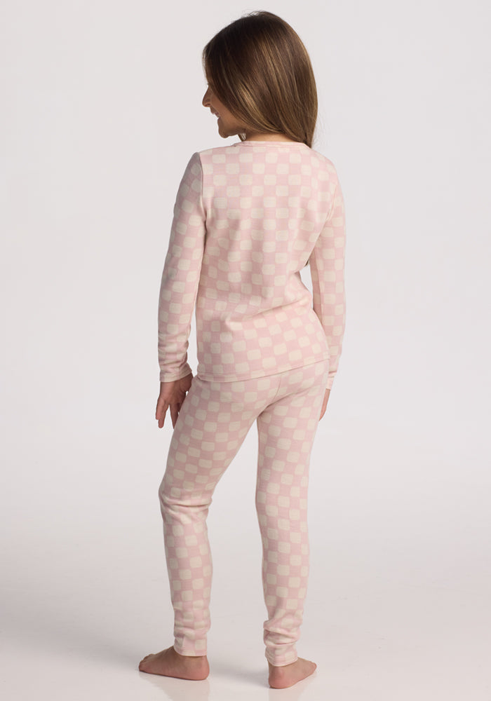 A young girl with long brown hair stands barefoot on a white background, wearing Woolx Kids Explorer Base Layer Bottoms in Hopscotch Pink check, layered under pink and white pajamas.