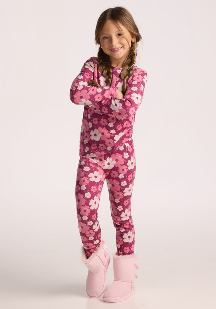 A young girl with braided hair, arms crossed and smiling, wears a pink floral pajama set over Woolx Kids Explorer Base Layer Bottoms - Rosie Posie and light pink fuzzy slippers. The background is plain white.