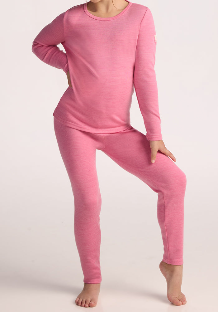 A child stands barefoot against a plain light background, wearing Woolx Kids Explorer Base Layer Bottoms in Pinky Promise and a matching long-sleeve top, with one hand on their hip and one leg slightly bent.