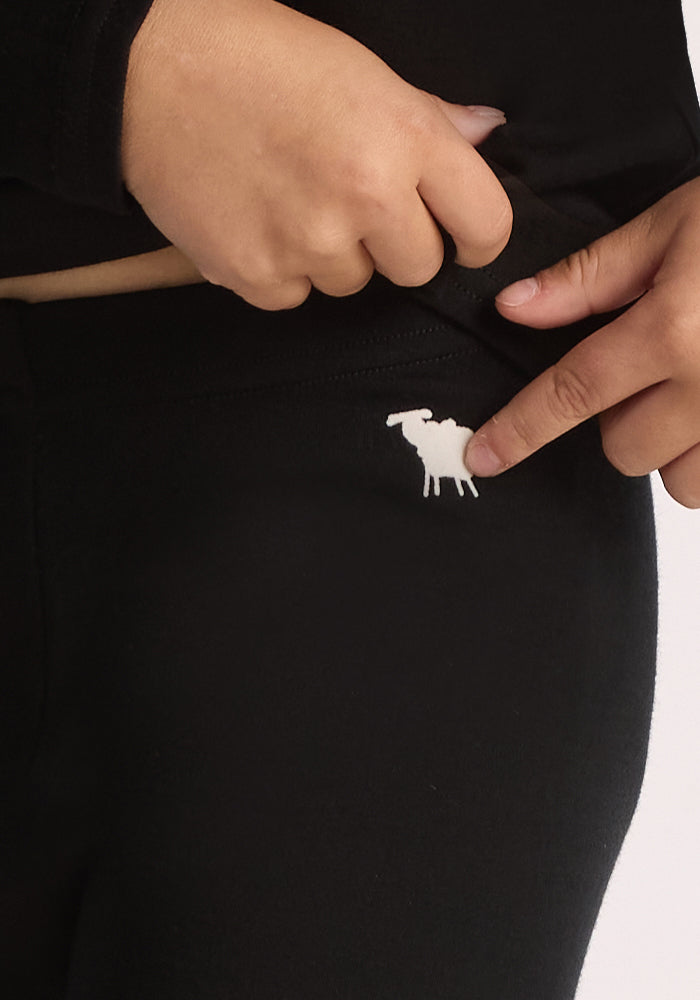 A person points to a small white moose logo on the waistband of Woolx Kids Explorer Base Layer Bottoms in black, while their other hand lifts the shirt slightly to reveal the logo.
