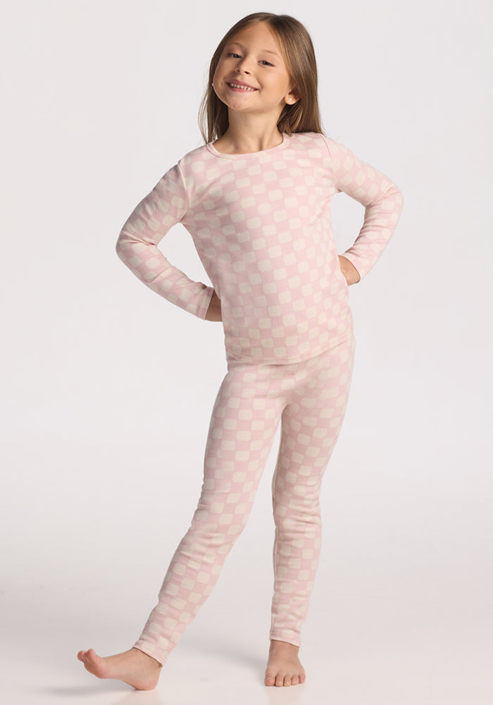 A young girl with long hair stands barefoot, smiling with her hands on her hips, wearing Woolx Kids Explorer Base Layer Bottoms in Hopscotch Pink against a plain white background.
